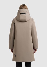 Beige lang frakke med høj krave og stor hætte, lavet af glat materiale. Inkluderer et logo-patch nær sømmen og lige skårne ærmer.