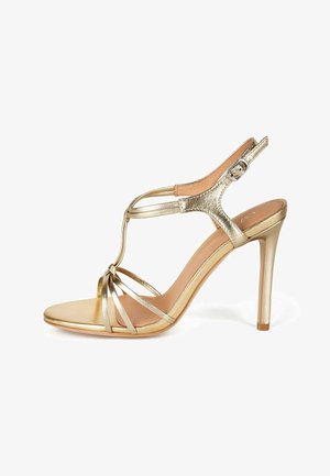 Sandalias de tacón alto doradas con tiras, diseño con tira T, acabado metálico y hebilla en el tobillo ajustable. Presenta un upper y suela lisos y texturizados.