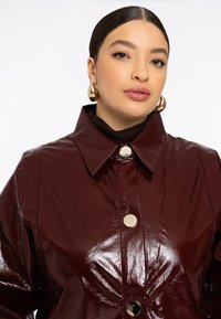 Femme avec les cheveux plaqués en arrière portant de grandes boucles d'oreilles en or et un manteau rouge foncé brillant boutonné avec un large col.