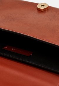 Brązowa torebka kopertowa z materiału przypominającego skórę, z metalowym zapięciem, o gładkiej fakturze i czarnym wykończeniu wnętrza. Widoczna metka wewnętrzna.