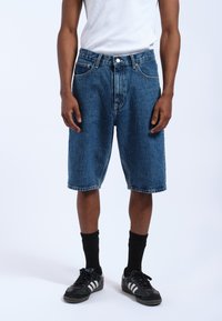 Pantalones cortos de mezclilla en azul oscuro con un corte holgado, que cuentan con cinco bolsillos, trabillas para el cinturón y un dobladillo recto. Llevados con una camiseta sin mangas blanca y zapatillas negras.
