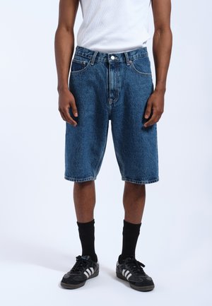 Denim shorts in dark blue with a loose fit, featuring five pockets, belt loops, and a straight hem. Worn with a white tank top and black sneakers.