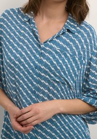 Femme portant une chemise bleue à boutons avec un motif abstrait blanc, les bras croisés légèrement devant, mettant en valeur le col et la poitrine.