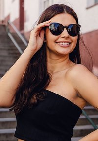 Černé sluneční brýle ve tvaru kočičích očí s reflexními skly. Modelka má na sobě černý top bez ramínek a stojí na betonových schodech. Dlouhé vlnité vlasy.