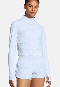 Mujer con una camiseta deportiva de manga larga y corte cropped de color azul claro con agujeros para los pulgares y pantalones cortos de running de cintura alta a juego, contra un fondo blanco.