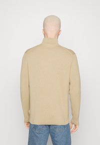 Maglione beige a coste con colletto alto, maniche lunghe e texture lavorata a maglia; presenta accenti rinforzati sulle spalle. Indossato con jeans blu.