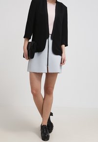 Kobieta w czarnym żakiecie, jasnoróżowej bluzce, jasnoniebieskiej spódnicy z zamkiem z przodu, czarnej kopertówce i czarnych, masywnych butach na platformie.