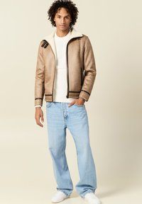 Giacca in suede marrone con colletto in pelliccia sintetica bianca, chiusura zip frontale, due tasche laterali e rifiniture scure, abbinata a jeans blu chiaro e sneakers bianche.