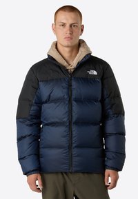 Blue puffer jacket with black upper panels, zip front, elastic cuffs, and logo on the chest. Material appears smooth and insulated.