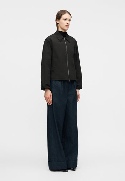 Γυναίκα όρθια, φοράει μαύρο μπουφάν με φερμουάρ πάνω από μαύρο τοπ, σκούρο μπλε παντελόνι με φαρδιά μπατζάκια και μαύρα μυτερά παπούτσια, με φόντο μια απλή επιφάνεια.