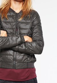 Vadderad bomberjacka i mörkgrått, med dragkedja framtill, ribbade muddar och en texturerad midja med en slät, glansig yta.