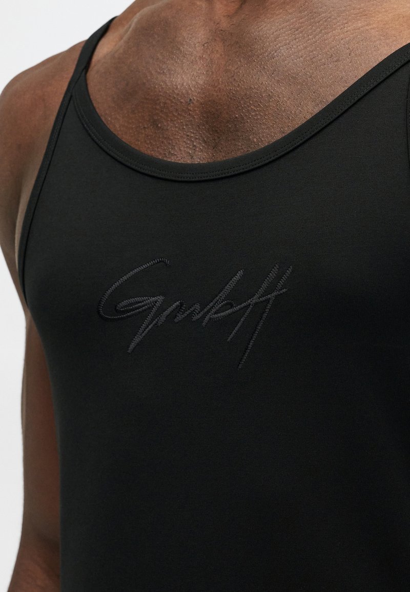 Primo piano di una persona che indossa una maglietta nera senza maniche con un piccolo logo ricamato "Gmlkft" sul petto.