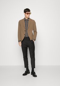 Bruine op maat gemaakte blazer over een geruite shirt, slim fit zwarte broek en zwarte schoenen. Accessoires omvatten bruin zonnebril. Glad texturen door het geheel.