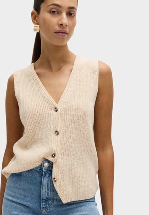 Kvinde iført ærmeløs beige strikket vest med knapper, stoppet ned i blå denimjeans med guld geometriske øreringe, neutral baggrund.