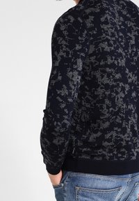 Marineblauer Strickpullover mit einem strukturierten Muster in Grautönen, gerippten Bündchen und Saum, figurnahem Schnitt und einem Reißverschluss-Detail an der Seite.