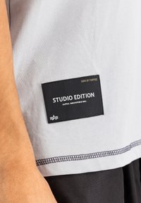 Světle šedé tričko s černou etiketou, na které je napsáno "STUDIO EDITION" a "ALPHA INDUSTRIES INC." s kontrastním černým švem.