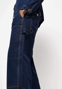 Mörka denim overaller med en avslappnad passform, med sidofickor, kontrasterande sömmar och knappstängningar på axlarna och ärmsluten.