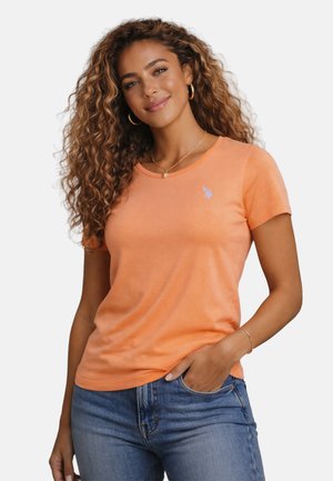 Woman with curly hair wearing an orange short-sleeve t-shirt and blue jeans, with one hand in pocket, smiling at the camera.