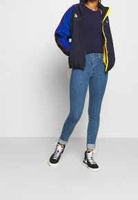 Cazadora azul marino con acentos coloridos, jeans oscuros ajustados y zapatillas negras de caña alta. Presenta puños elásticos y cierre de cremallera.