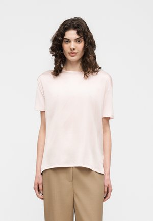 Tânără cu păr creț și închis la culoare, purtând un tricou roz pal cu mâneci scurte și pantaloni largi bej, stând în fața unui fundal simplu.