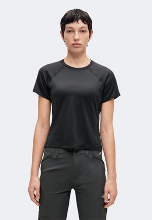 Personne debout portant un t-shirt sombre ajusté à manches courtes et un pantalon sombre avec des poches zippées sur un fond clair uni.