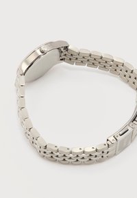 Ασημένιο μεταλλικό ρολόι με συνδεδεμένο βραχιόλι και κλειστό κούμπωμα, εμφανίζεται από πίσω και πλάγια σε απλό λευκό φόντο.