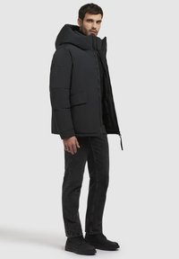 Muškarac u crnoj jastučićastoj jakni s kapuljačom, tamnim trapericama i crnim čizmama stoji naslonjen na jednostavnu svijetlu pozadinu.