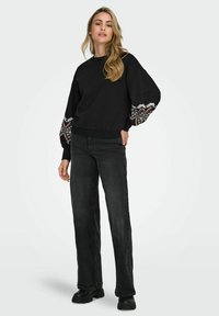 Črna trenerka z vzorčenimi vezenimi manšetami, okroglim izrezom in sproščenim krojem. Ujemite jo z visokim pasom črnimi kavbojkami in masivnimi črnimi škornji.