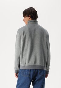 Sudadera de forro polar gris con cuello alto, puños y dobladillo acanalados. Textura suave, corte relajado, combinada con pantalones de mezclilla azules.