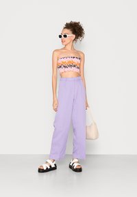 Top cropped sem alças em várias cores com padrões geométricos, combinado com calças largas em lavanda claro, sandálias pretas com alças brancas e uma bolsa bege.