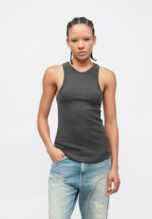 Jeune femme aux cheveux tressés portant un débardeur côtelé gris foncé et un jean clair déchiré, une main dans la poche, fond neutre.