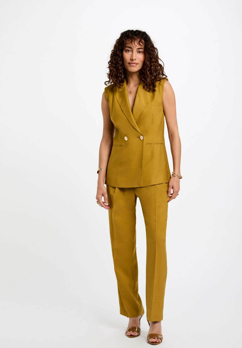 Chaleco a medida de color mostaza con diseño de doble botonadura y pantalones rectos a juego. Tejido suave, estilo minimalista, sin mangas.