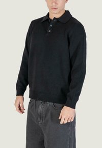 Maglione nero con colletto, tre bottoni, polsini e orlo a coste. Texture morbida, indossato sopra jeans grigi.