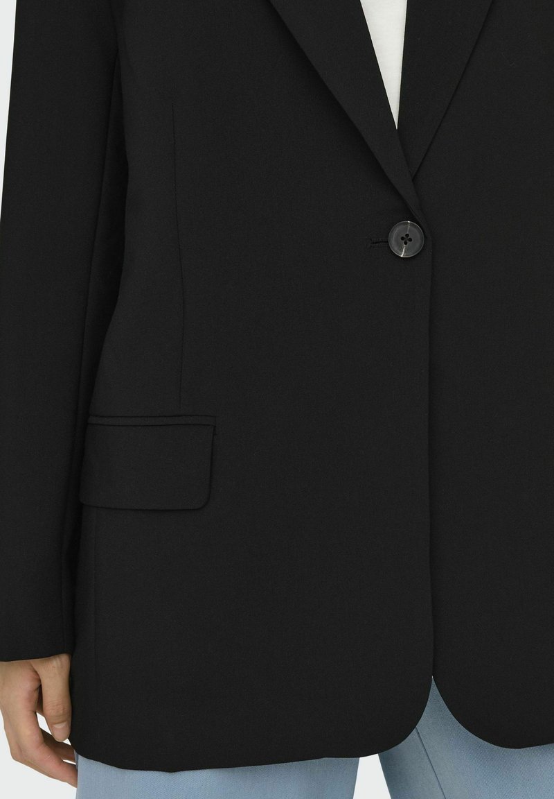 Schwarzer Blazer aus strukturiertem Stoff, mit einem Ein-Knopf-Verschluss, Reverskragen und einer Seitentasche. Kombiniert mit hellblauen Jeans.
