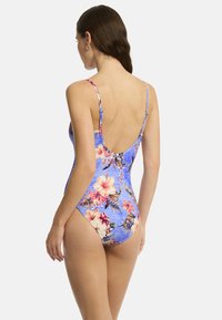 Modèle présenté portant un maillot de bain une pièce à fleurs bleues avec un dos décolleté et des fines bretelles, mettant en valeur de vibrantes fleurs d'hibiscus roses.