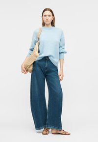 Γυναίκα που φοράει ανοιχτό γαλάζιο πουλόβερ, τζιν παντελόνι με φαρδιά πόδια, μπεζ σανδάλια και κρατάει πλεκτή μπεζ τσάντα ώμου μπροστά σε μονόχρωμο φόντο.