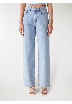 Kobieta w jasnoniebieskich jeansach z szerokimi nogawkami i białych sandałach z paskami i odkrytymi palcami, stojąca na jednolitym białym tle.