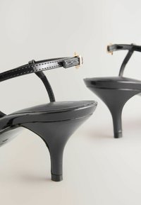 Sorte patenterede højhælede sandaler med tynde stropper, guld hardware detaljer og en slank spids tå. Skinnende overflade og buet hæl.