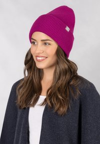 Paarse geribde gebreide beanie met een omgeslagen rand, voorzien van een klein logo-opslag. Draag het met een donkergrijze cardigan over een wit shirt.