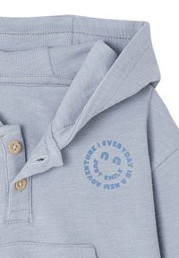 Helle blaue gestrickte Babykleidung mit Kragen, Holzknöpfen und blauer Smiley-Grafik mit kreisförmigem Text auf der Brust.