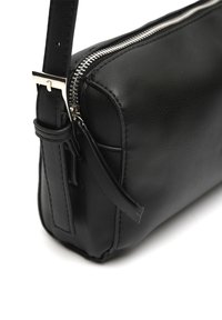 Angolo di una borsa a tracolla in pelle nera con zip silver e fibbia per tracolla regolabile su sfondo bianco.