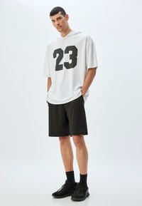 Hvid oversized hættetrøje med sort mesh nummer "23," parret med sorte shorts og sorte sportssko. Simpelt, moderne design.