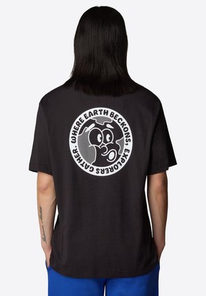Personne aux cheveux raides et foncés porte un t-shirt noir avec un graphique circulaire blanc de la Terre avec un visage de dessin animé et le texte « Là où la Terre appelle, les explorateurs se rassemblent. »