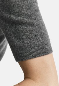 Nahaufnahme eines grauen gestrickten Ärmels, der über dem Ellenbogen endet und die feine Stoffstruktur sowie ein dezentes Logomuster zeigt, das in das Material eingewebt ist.