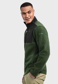 Dunkelgrüne Fleecejacke mit einem schwarzen oberen Teil, einem Reißverschluss vorne und elastischen Bündchen. Auffälliges Markenlogo auf der Brust.