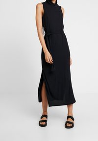 Czarna długa sukienka na ramiączkach z wysokim kołnierzem, bocznymi rozcięciami i paskiem w talii. Gładka tkanina o lekkim po�łysku.