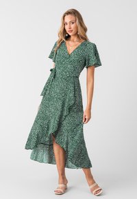 Vestido verde con lunares blancos, mangas cortas con vuelo y un dobladillo con volantes. Tejido suave con un detalle de lazo en la cintura.
