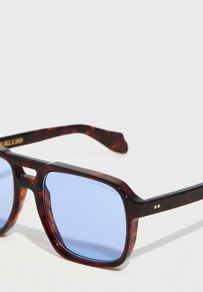 Γυαλιά ηλίου με τετράγωνους σκελετούς από καβούκι χελώνας, φακούς με μπλε απόχρωση και διακριτικές λεπτομέρειες με χρυσά αστέρια στα πλαινά μέρη.