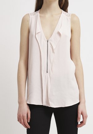 Sleeveless light pink blouse with front zipper, lace details on shoulders, worn with black pants by a standing person against a plain background.