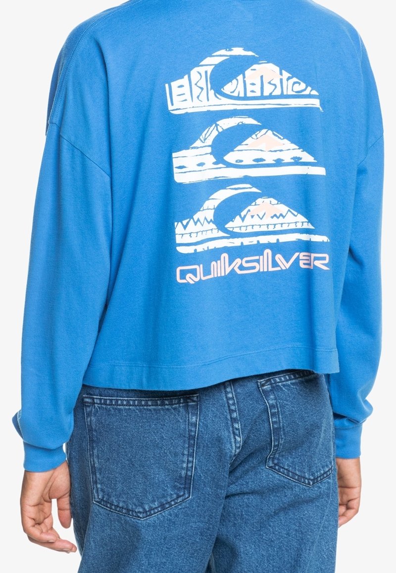 Niebieska, długorękawowa, krótka bluza z graficznym motywem fal i gór w bieli z różowymi akcentami, zestawiona z dżinsami.